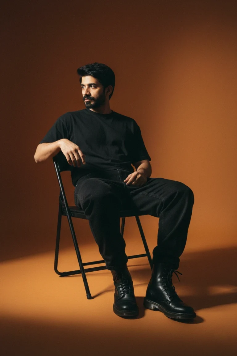 A man in a black t-shirt and dark jeans sitting on a folding chair against an orange studio backdrop, showcasing minimalist male studio portrait photography.